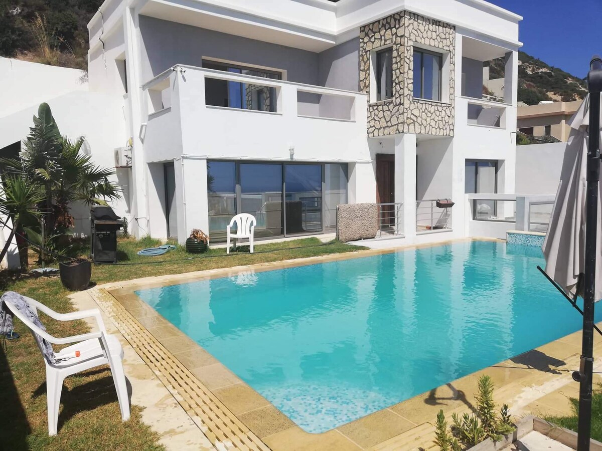 A modern house features a sleek, rectangular swimming pool surrounded by a grassy area. Expansive glass windows reflect the bright exterior and allow views of the serene landscape. A few lounge chairs are positioned beside the pool, offering a relaxing space for guests.
