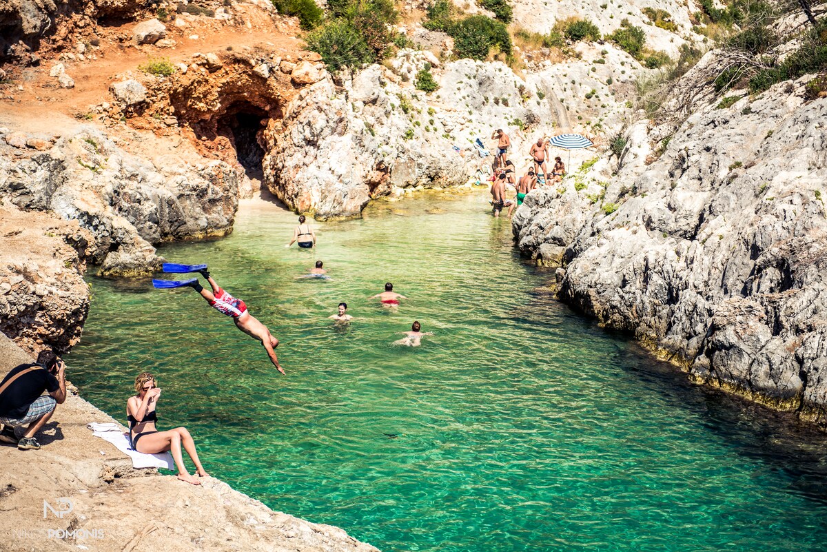 A rocky cove is depicted, featuring clear turquoise waters. Several individuals are seen swimming or lounging on the rocks. One person is in mid-air, diving into the water, while another is seated on the edge, enjoying the sun. Lush greenery is visible in the background.