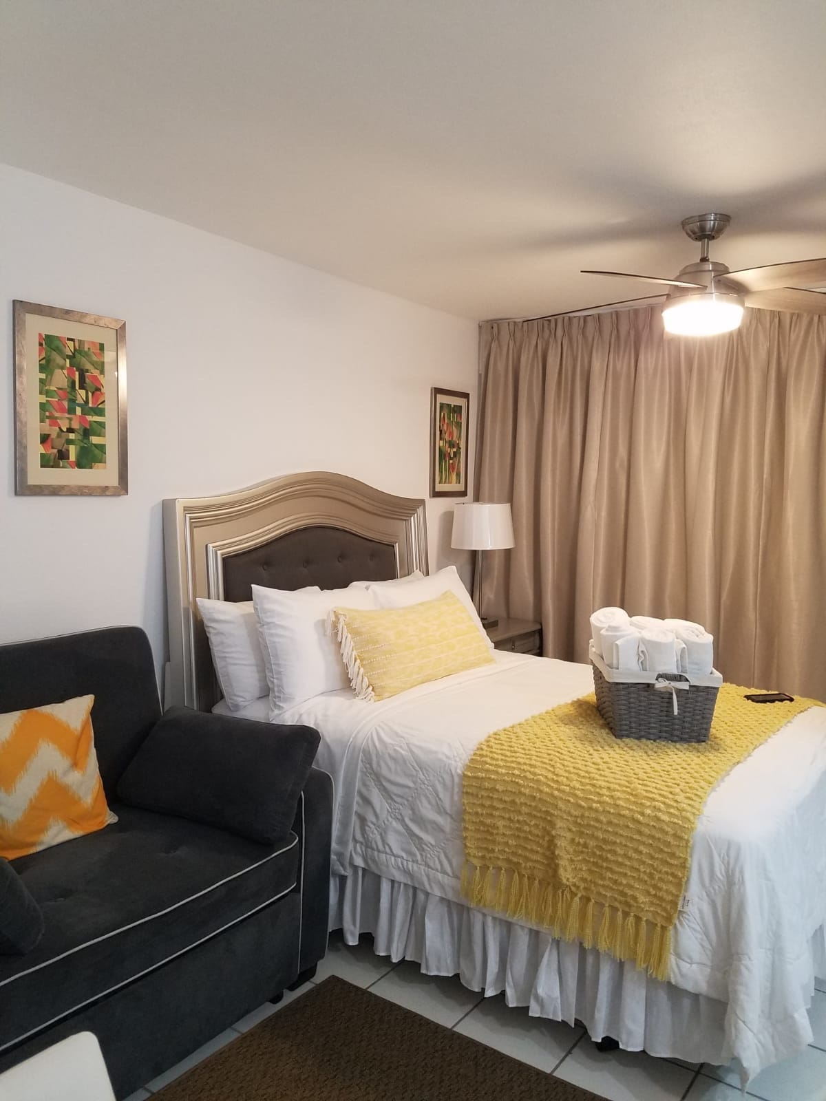 A cozy sleeping area features a spacious bed dressed in white linens accented with a yellow throw. A gray armchair sits nearby, and artwork adorns the walls. Soft beige curtains frame the window, with a ceiling fan providing a gentle light.