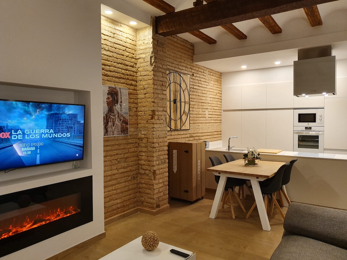 An open-plan living area is presented, featuring a fireplace with a warm glow beneath a modern television. A light wood dining table with matching chairs occupies the center, while a sleek kitchen with minimalist cabinetry and appliances is visible in the background against a textured brick wall.