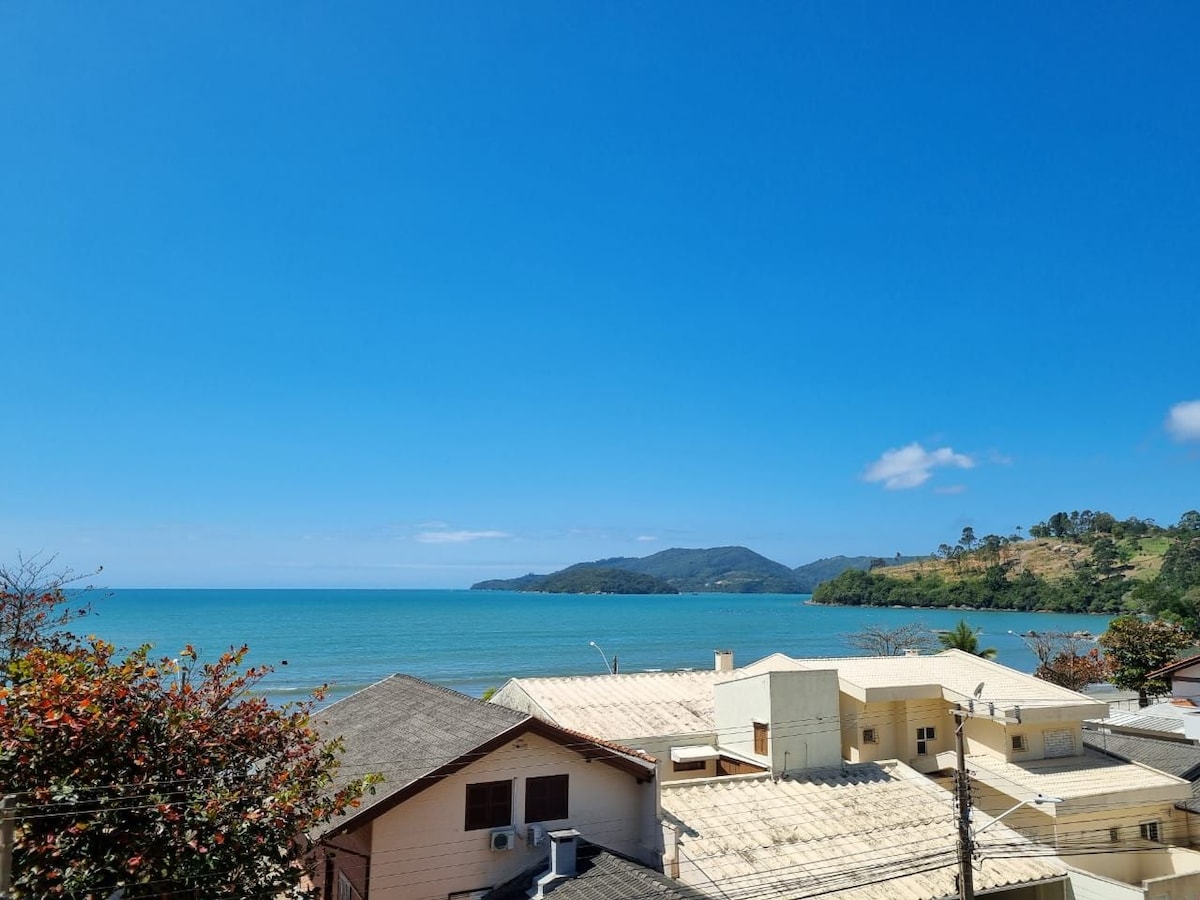 A serene coastal view showcases calm blue waters meeting a clear sky. Houses with varied rooftops are visible in the foreground, while distant hills and trees frame the horizon, contributing to the peaceful scenery.