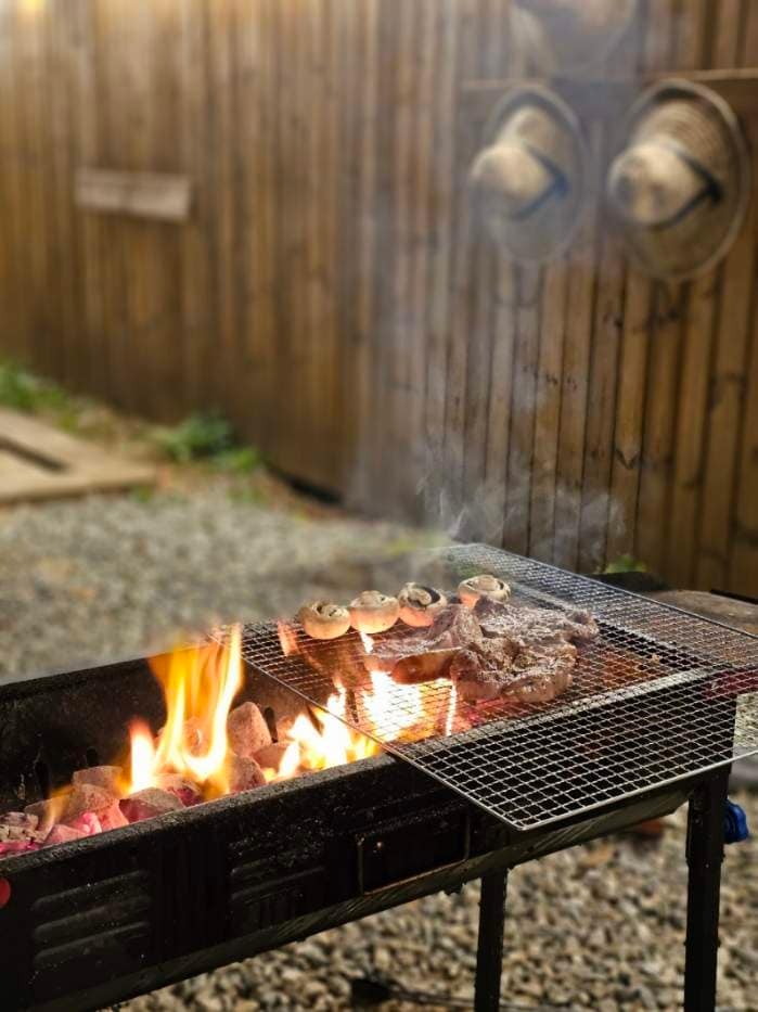 A charcoal grill is positioned outdoors, with steaks and mushrooms cooking over an open flame. Smoky wisps curl upwards against a wooden backdrop, while a gravel surface lays underneath the setup. Two straw hats hang on the fence, adding a rustic element to the scene.