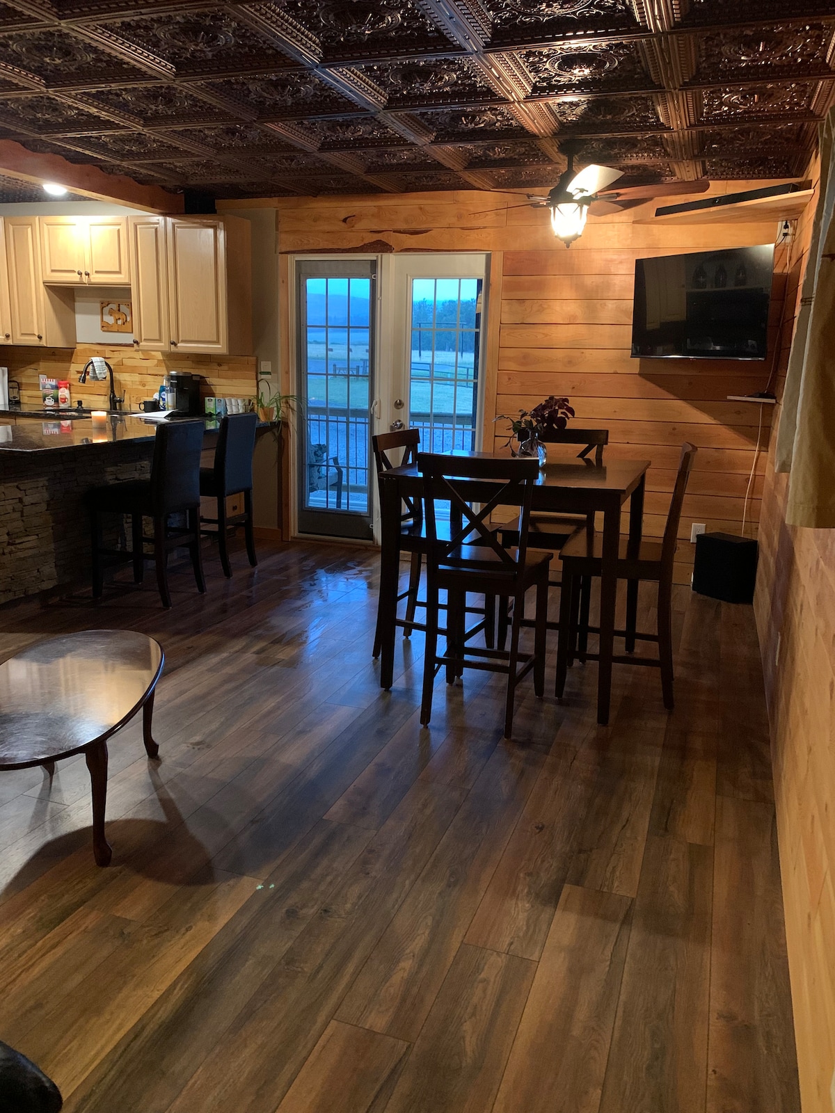 The open living area features a dining table surrounded by dark wooden chairs, with a cozy seating area nearby. A modern kitchen is visible, showcasing light cabinetry and appliances. Natural light enters through sliding glass doors, leading to an outdoor view.