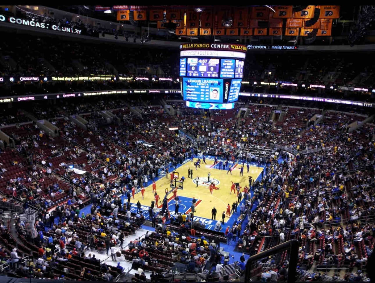 A wide view of an indoor sports arena packed with spectators, highlighting the basketball court at center stage. Large screens hang from the ceiling, displaying scores and game information. Bright lights illuminate the court, and players can be seen warming up before the game.