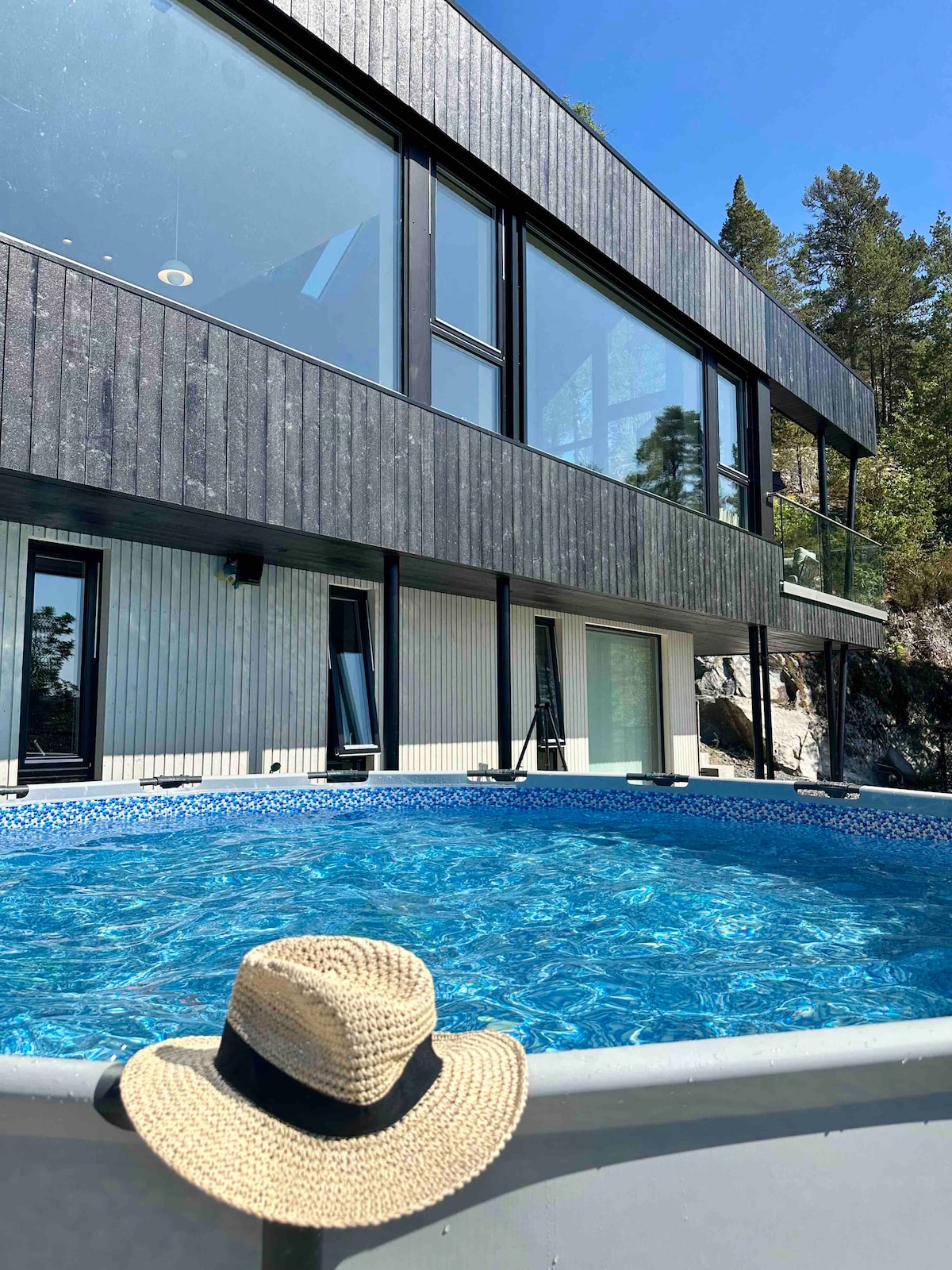 A bright blue above-ground pool is situated in the foreground, with a straw hat resting on its edge. A modern house is visible behind the pool, featuring large glass windows and wooden siding that blends with the natural surroundings.