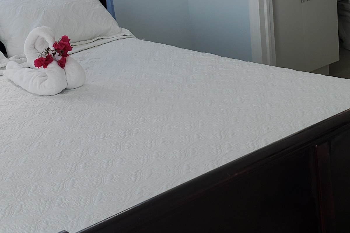 A spacious bed is dressed in a white, textured coverlet. Two rolled towels are arranged decoratively on the bed, accompanied by a small cluster of pink flowers. Soft light illuminates the area, creating a serene and inviting space.