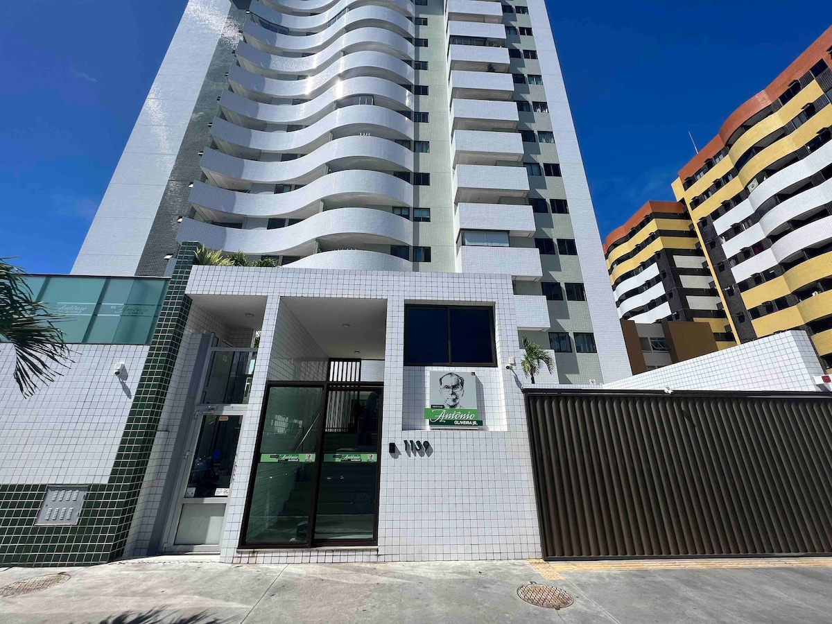 The exterior of a modern building is showcased, featuring a sleek design with large windows and a bright facade. Green accents are visible, along with a gated entrance. A clear blue sky provides a vibrant backdrop, highlighting the structure's height and architectural style.