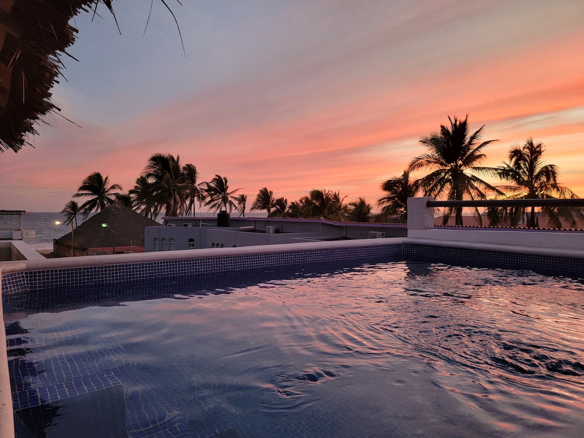 A tranquil jacuzzi is illuminated by the soft glow of a sunset, reflecting warm hues on the water's surface. Palm trees frame the view, while the ocean is visible in the background, enhancing the peaceful ambiance of the terrace.