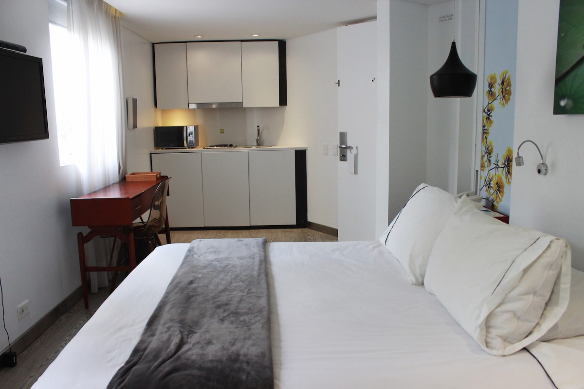 A spacious bedroom area includes a large bed with plush white bedding, complemented by a soft gray throw. A compact kitchenette is visible in the background, featuring modern cabinetry and appliances. A desk with a chair sits nearby, and natural light enters through a large window.