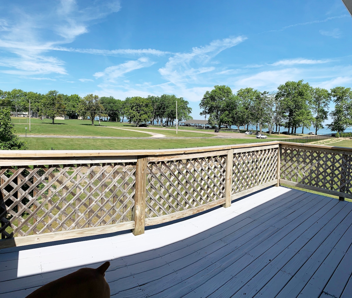 A spacious outdoor deck is visible, featuring wooden railings and a well-maintained white floor. The view showcases a grassy area extending towards trees and Lake Erie in the distance, under a bright blue sky with scattered clouds.