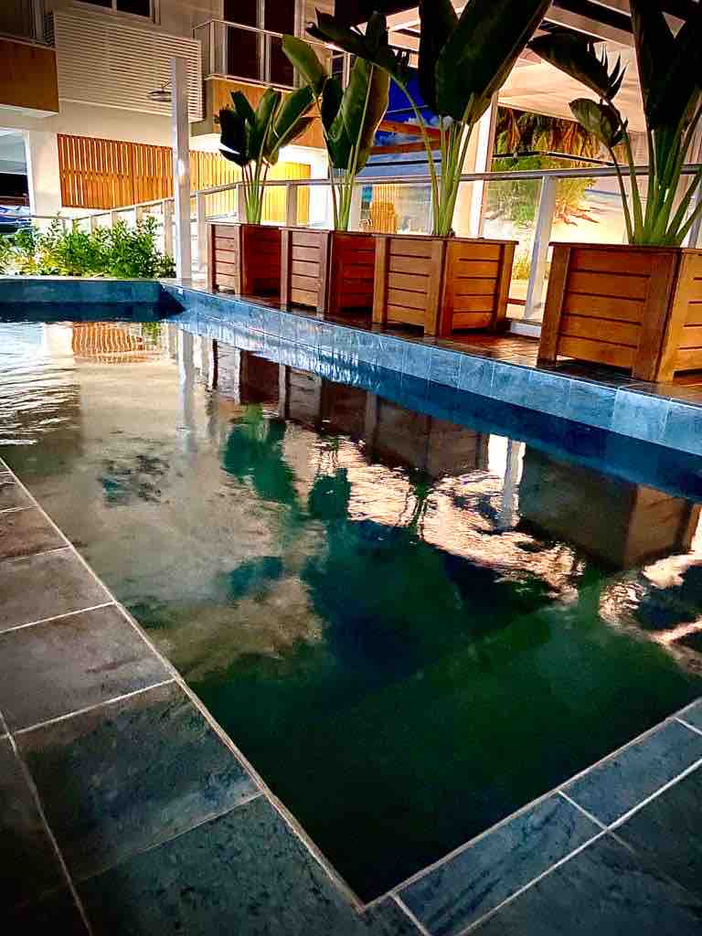 A tranquil pool area is framed by wooden planter boxes filled with lush green plants. Reflections of the surrounding space shimmer on the water's surface, creating a serene ambiance. The pool's tiled edges offer a smooth finish, enhancing the overall inviting environment.