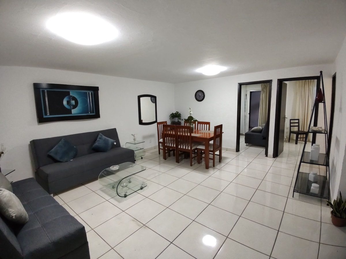 A comfortable living area features a gray sofa and a glass coffee table. A wooden dining table is surrounded by chairs, ideal for group meals. A mirror and artwork adorn the walls, and light fixtures illuminate the space, creating a bright and welcoming atmosphere.