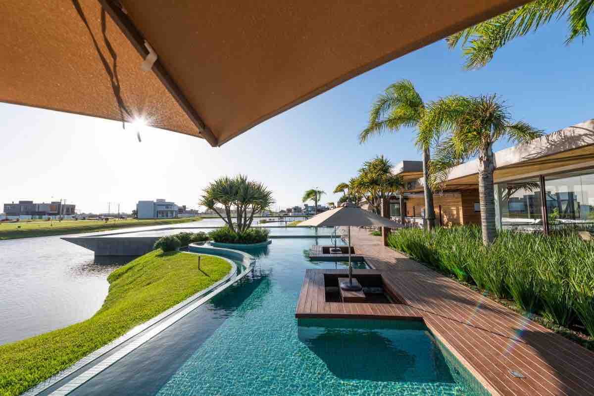 A serene outdoor area features a pool that extends alongside the lush greenery, leading to a picturesque view of the lake. Lounge chairs are positioned under large umbrellas, providing comfortable seating options. Sunshine reflects off the calm water, enhancing the tranquil ambiance.