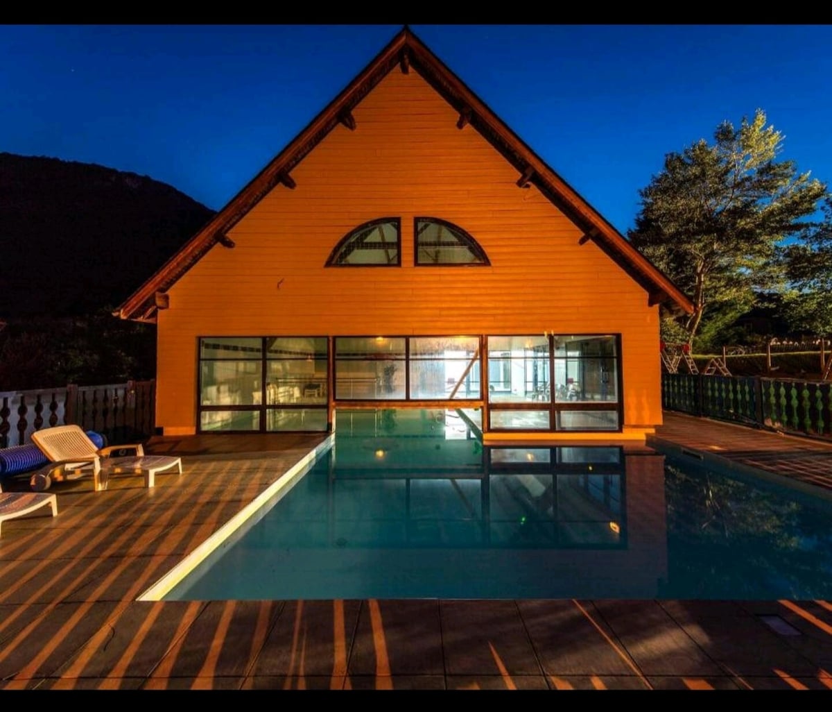 A brightly colored wooden structure features large windows that reflect the surrounding natural landscape. The swimming pool is illuminated, creating a serene ambiance. Lounge chairs are positioned nearby on a spacious deck, with shadows cast by the evening light adding depth to the scene.