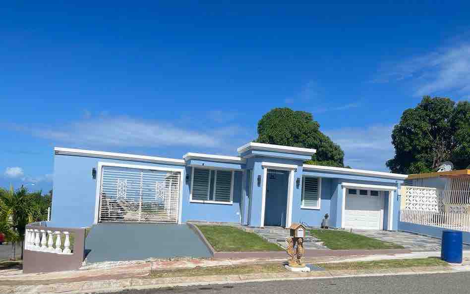 A modern light blue house features a streamlined design with large windows and a garage. The front yard includes a small landscaped area with grass and concrete pathways leading to the entrance. Lush trees are visible in the background under a clear blue sky.