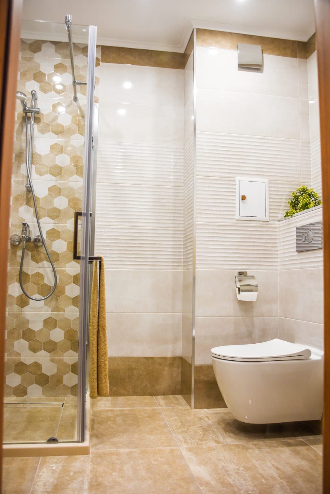 A modern bathroom is presented, featuring a spacious shower with a glass enclosure and a sleek showerhead. The walls are adorned with hexagonal tiles, complemented by earthy, neutral tones. A toilet is placed adjacent to a decorative plant, adding a touch of greenery to the space.