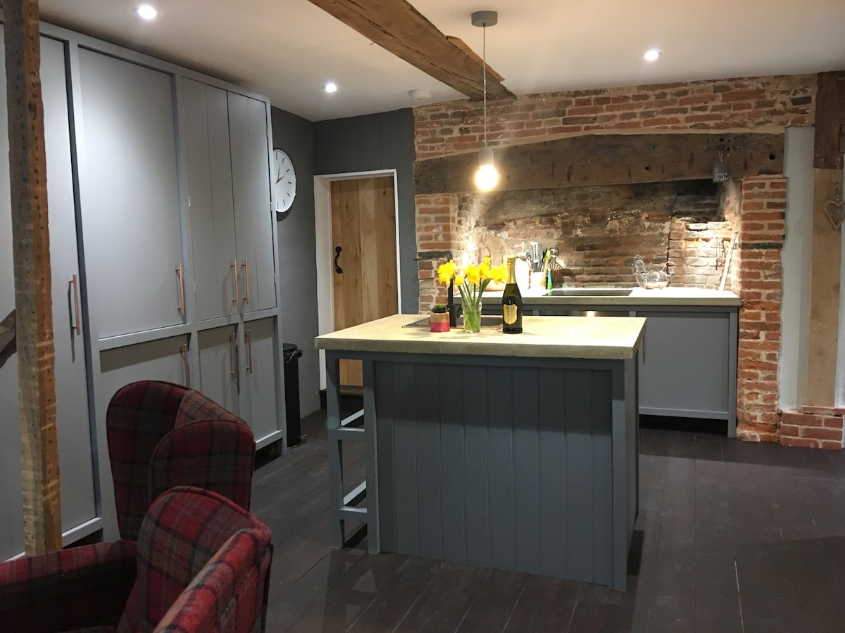 The kitchen features a large central island with a light countertop, complemented by modern cabinetry and exposed brick accents. A cozy seating area is visible with plaid armchairs. The room is well-lit, showcasing a combination of functional appliances and decorative elements.