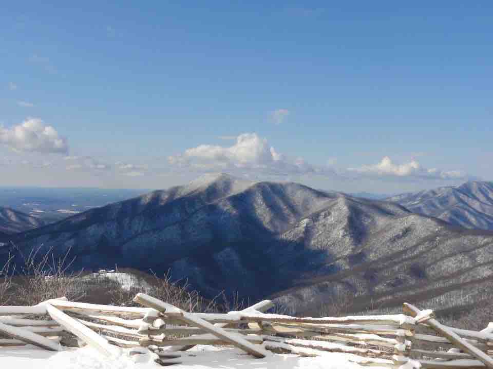 A panoramic view of snow-covered mountains is presented under a clear blue sky. The peaks are accented with patches of sunlight and distant clouds. A wooden fence runs along the foreground, adding a rustic element to the serene landscape.