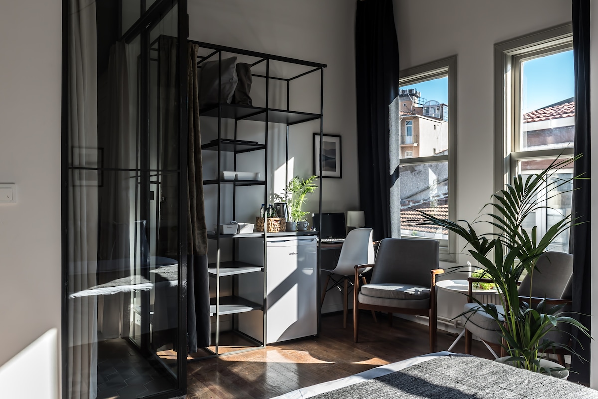 The minimalist studio features natural light streaming through two windows, illuminating a small seating area with two armchairs and a glass table. A shelving unit stands against a wall, while a small fridge is positioned nearby. Indoor plants add a touch of greenery to the space.