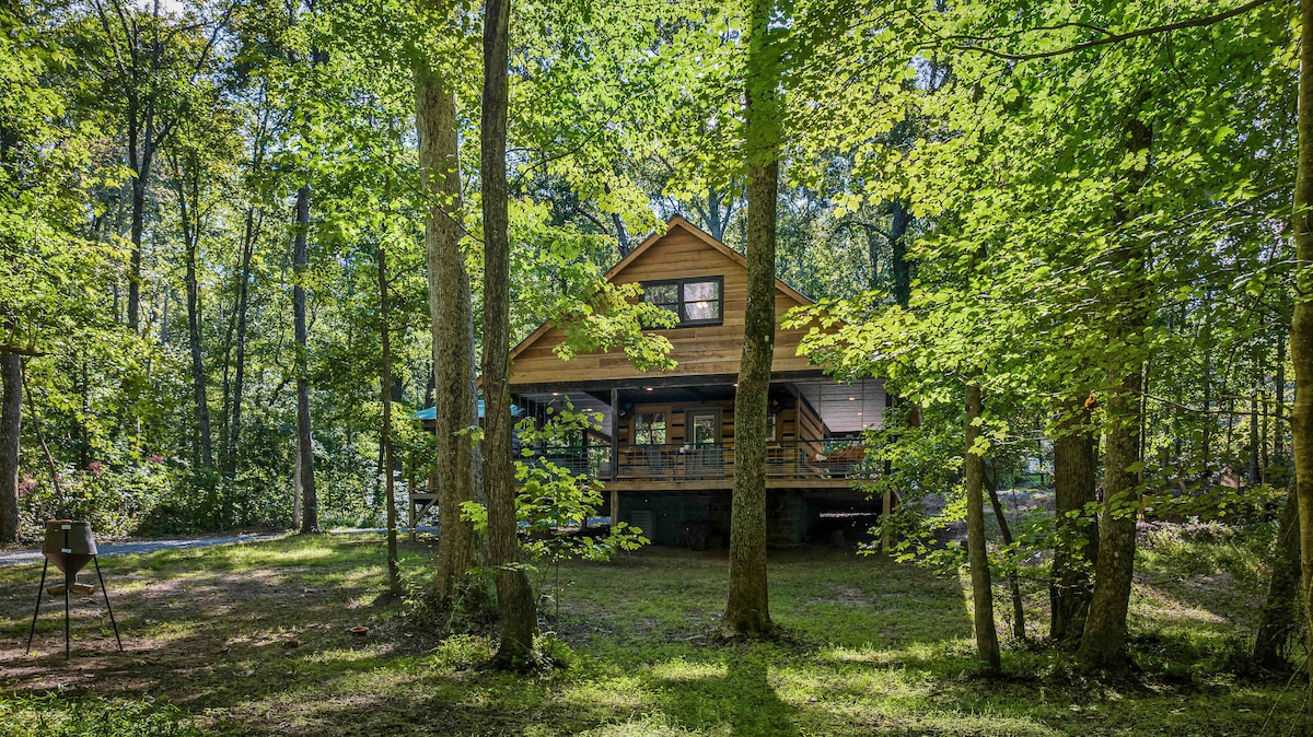 The custom cabin is nestled among tall trees, surrounded by lush greenery. Its wraparound porch offers shaded seating. Natural light filters through the leaves, creating a serene outdoor setting. An outdoor grilling area is visible nearby, enhancing the inviting atmosphere of the property.