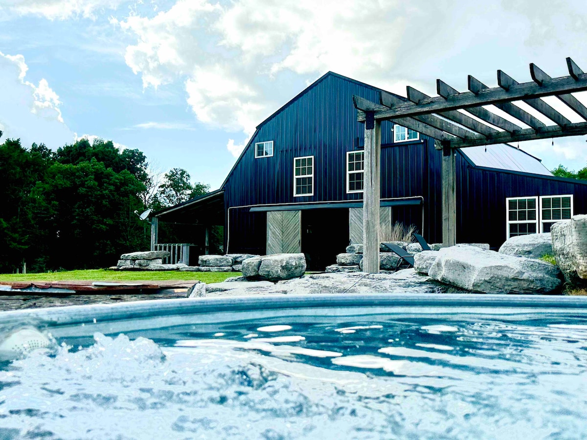 A stock tank pool sits in the foreground, with water rippling gently. In the background, a charming barndominium features dark blue siding and large windows. Stone landscaping surrounds the pool, complemented by a wooden pergola that enhances the rustic setting.