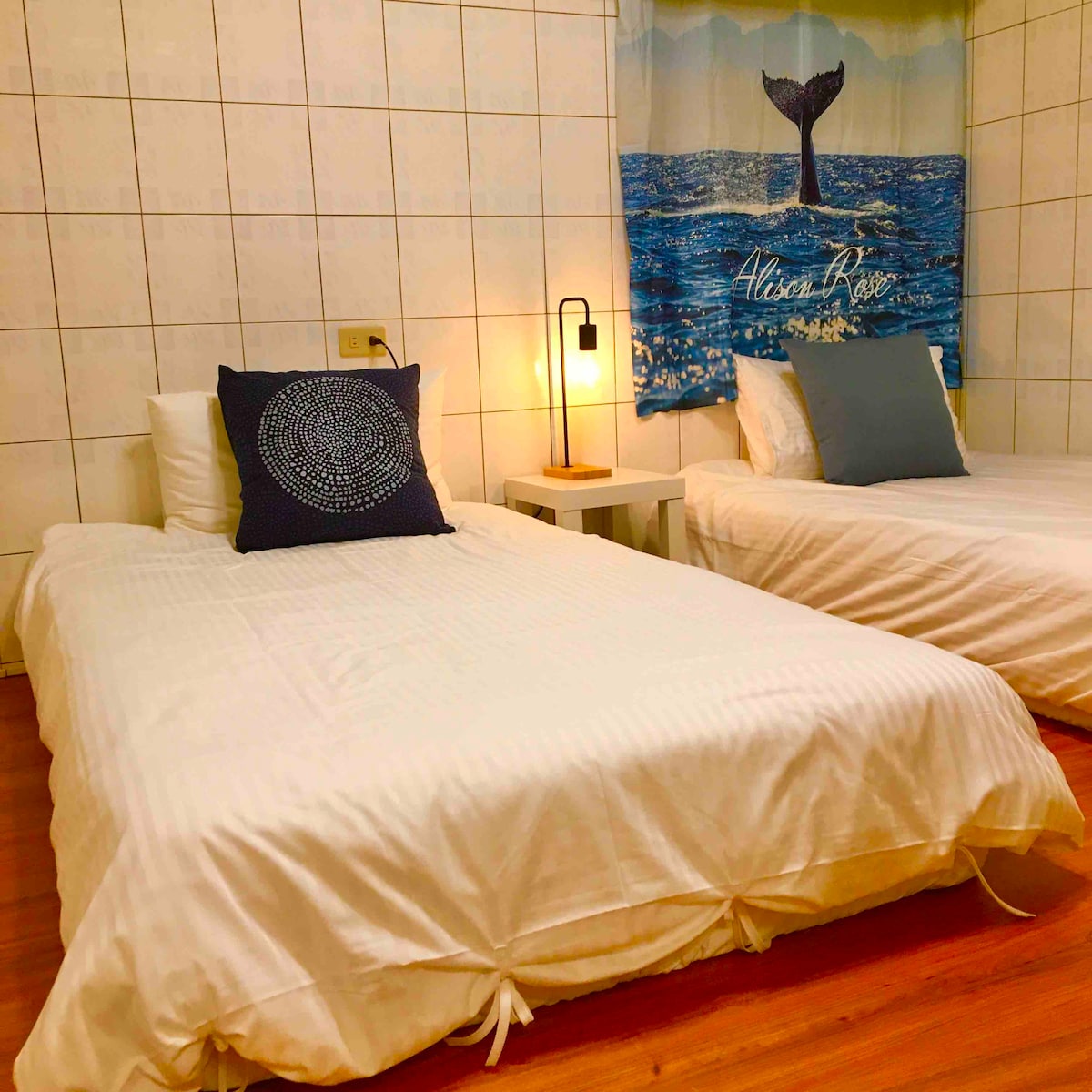 A tranquil sleeping area features two spacious beds with white linens, accented by pillows in calming colors. A bedside lamp stands next to one bed, providing soft illumination. The wall behind displays a decorative tapestry depicting a serene ocean scene.