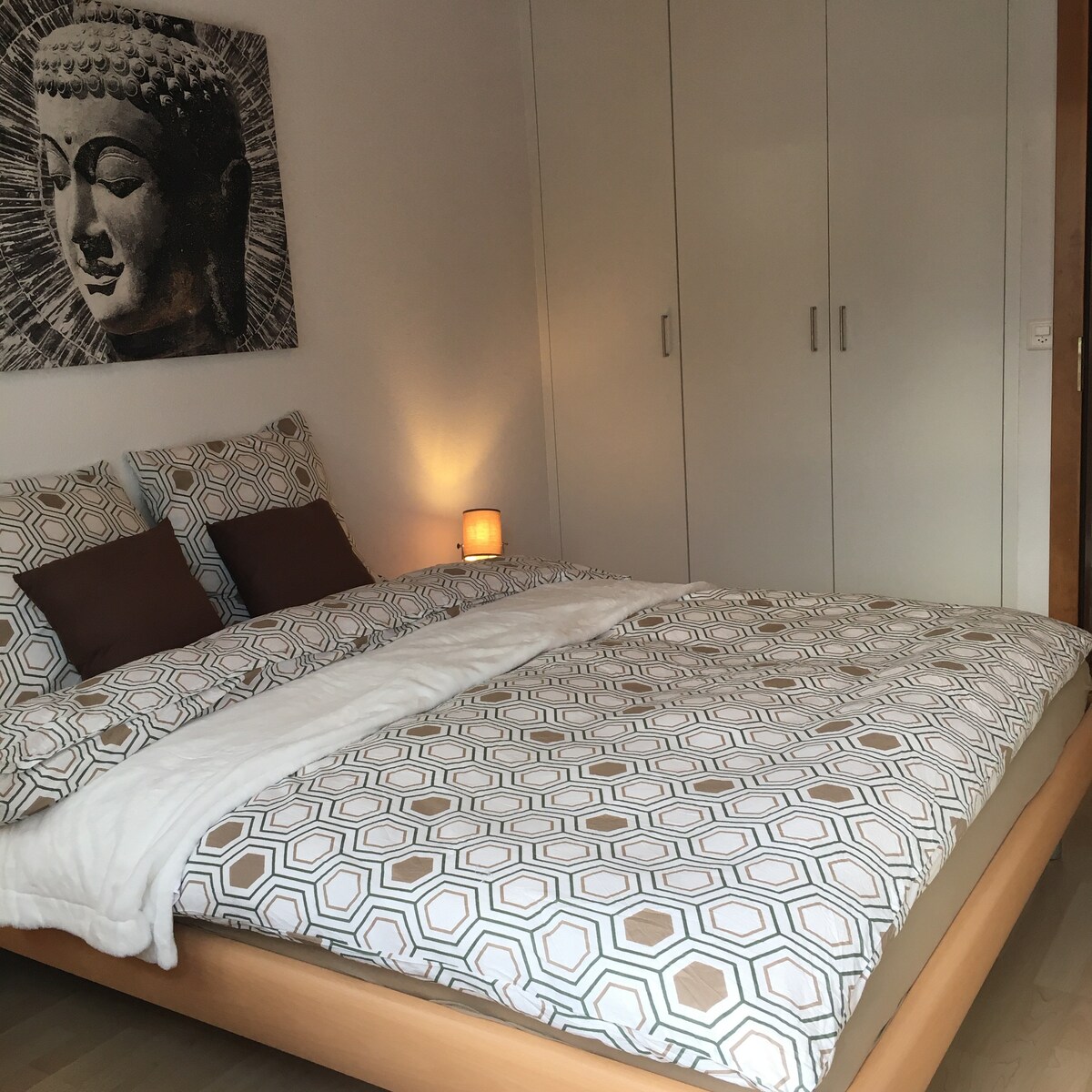A cozy bedroom features a spacious bed with a geometric patterned duvet cover and soft pillows. A bedside lamp provides a gentle glow, while a modern piece of art adorns the wall above the bed. Ample storage is available with built-in wardrobes.