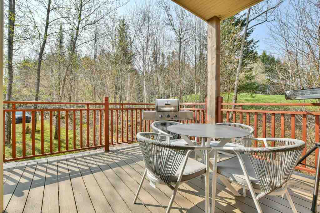 A wooden deck features a dining set with a round table and two chairs, positioned beside a barbecue grill. Surrounding trees provide a natural backdrop, while sunlight casts gentle shadows across the flooring.