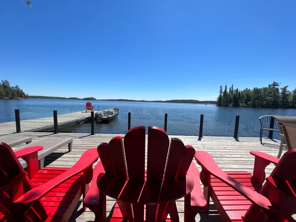 Bright red Adirondack chairs are arranged on a wooden deck overlooking a serene lake. A private dock extends into the water, where a boat is moored. The clear blue sky complements the natural surroundings of trees lining the shore.