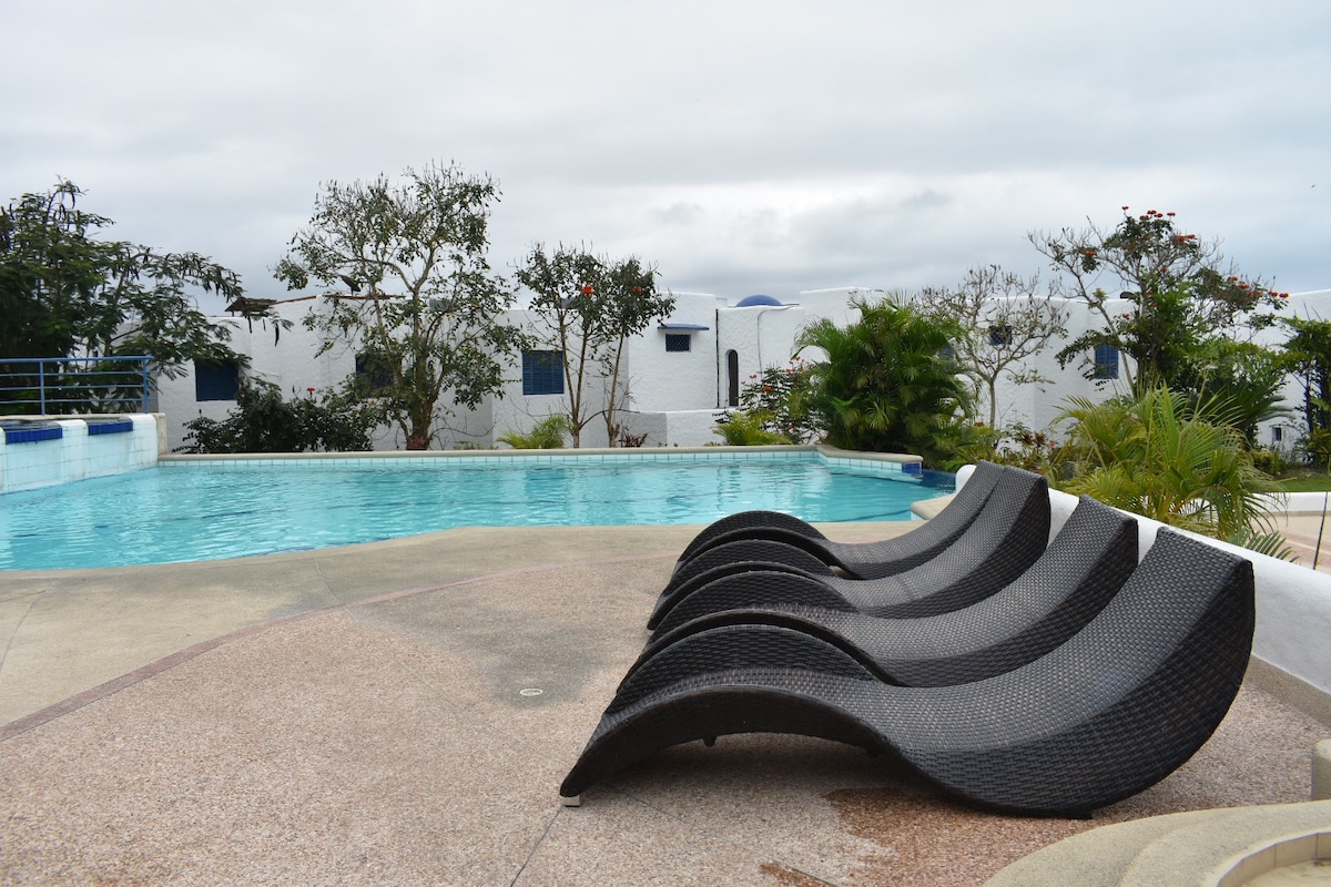A pool area is showcased with a clear blue pool surrounded by lounge chairs. Lush greenery is visible in the background, enhancing the outdoor space. The atmosphere appears tranquil, ideal for relaxation and sunbathing.