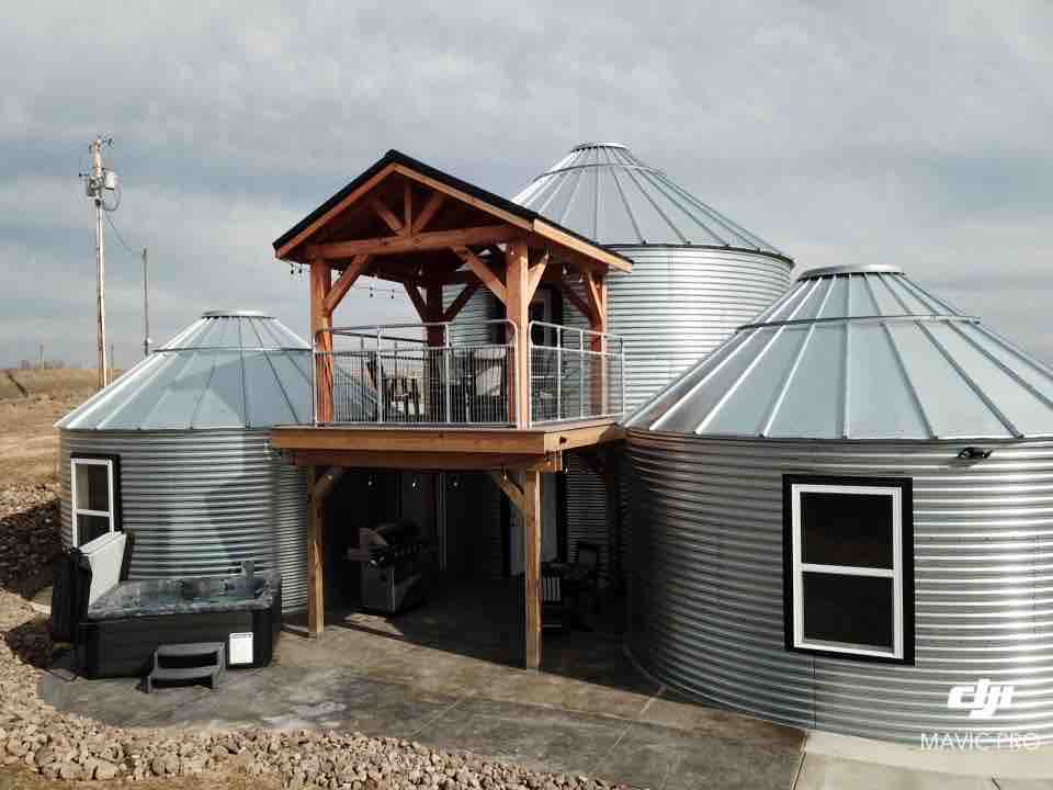 The unique grain bin structure features a combination of circular metal silos and a wooden deck. A hot tub is positioned on the lower level, while a balcony with railing provides additional outdoor space above. Large windows allow for natural light in the living areas.