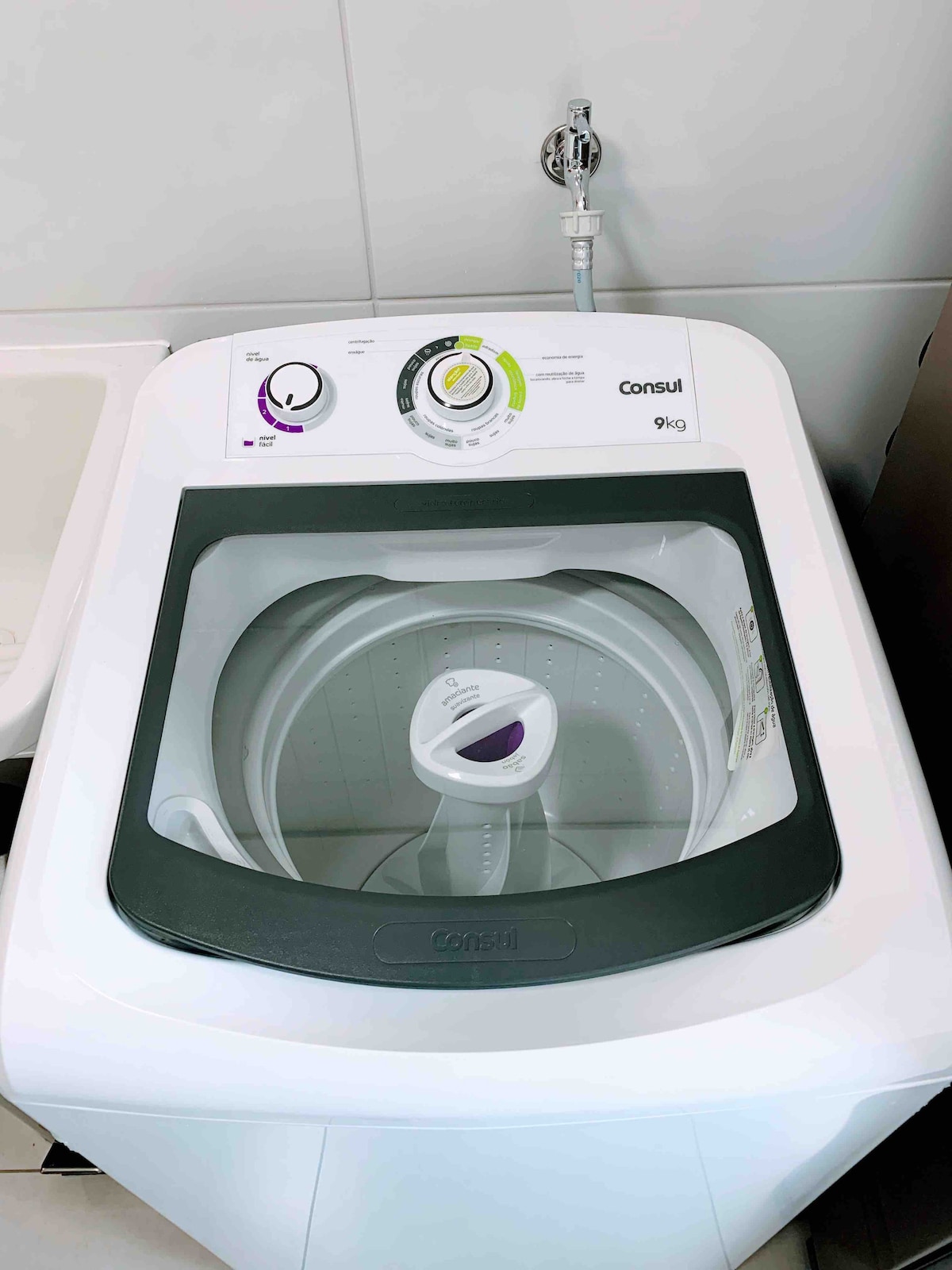A top-loading washing machine is displayed, featuring a modern design with a clear lid and control panel. The machine is designed for a 9 kg capacity and is positioned within a light-colored laundry area.