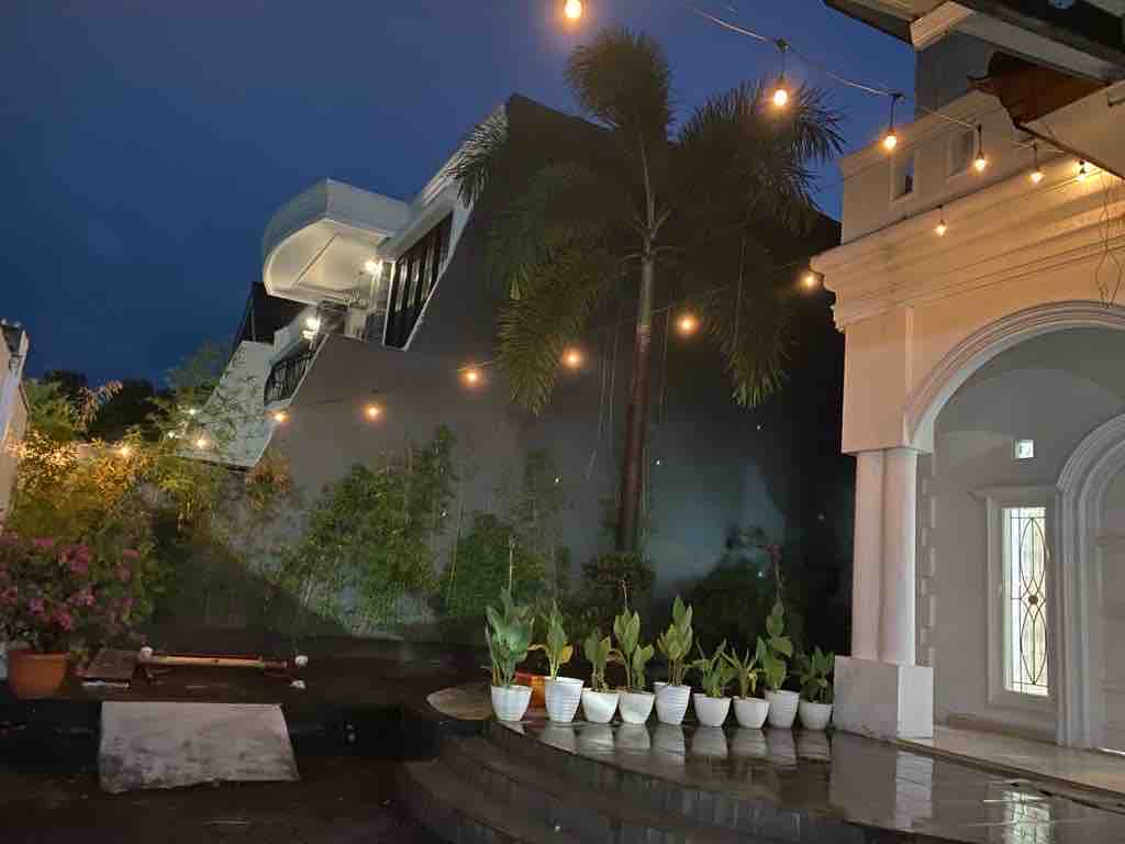The outdoor space features illuminated string lights draped over a charming patio area. Potted plants line a stone pathway, while lush greenery is visible in the background, creating a peaceful ambiance during the evening.