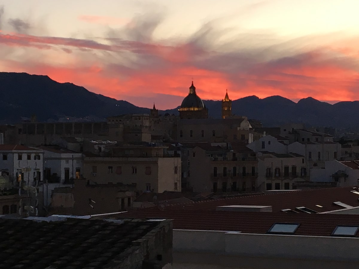 A sunset view displays a colorful sky with shades of orange and pink, framing the silhouette of buildings and mountains. The dome and towers of nearby structures are illuminated by the fading light, creating a striking contrast against the darkening landscape.