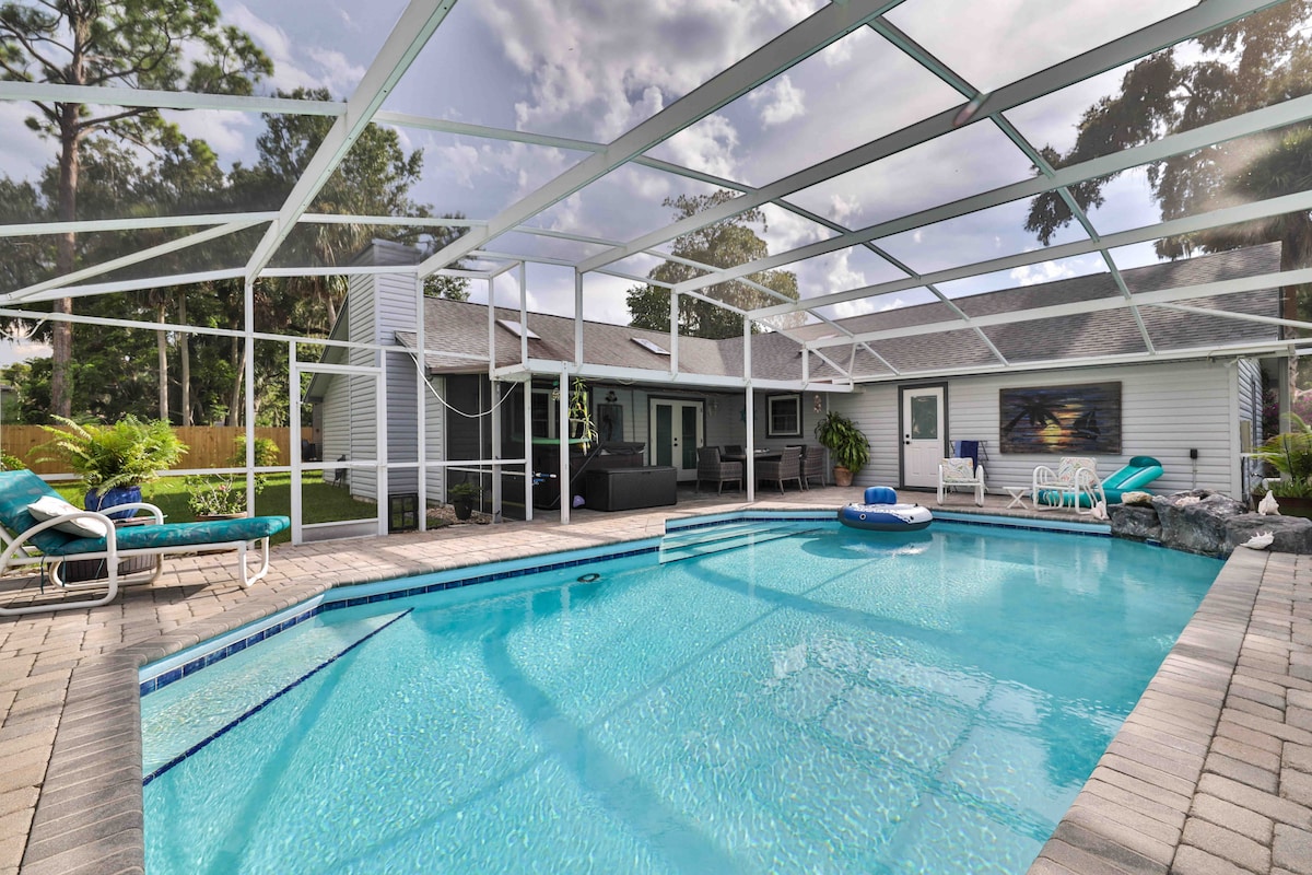 A custom-designed home features a serene outdoor area with a screened-in private pool. Comfortable lounge chairs are positioned around the pool, and a tranquil water feature enhances the setting. Lush greenery surrounds the area, providing a sense of privacy and relaxation.