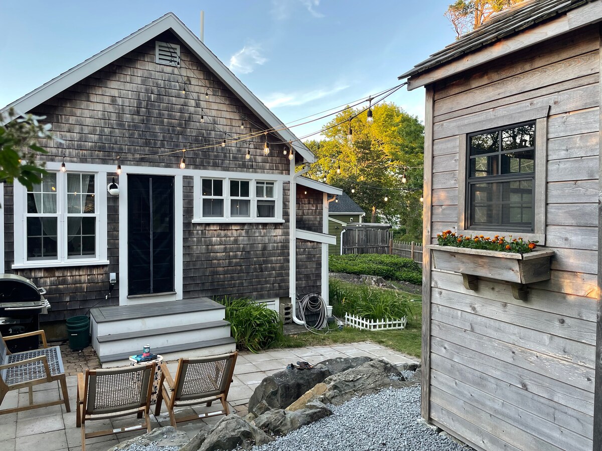 A charming exterior view of the cottage is presented, showcasing rustic shingle siding and white trim. A cozy patio area is highlighted, featuring wooden chairs and a small table. A shed is visible nearby, adorned with flowers in a window box, enhancing the outdoor appeal.