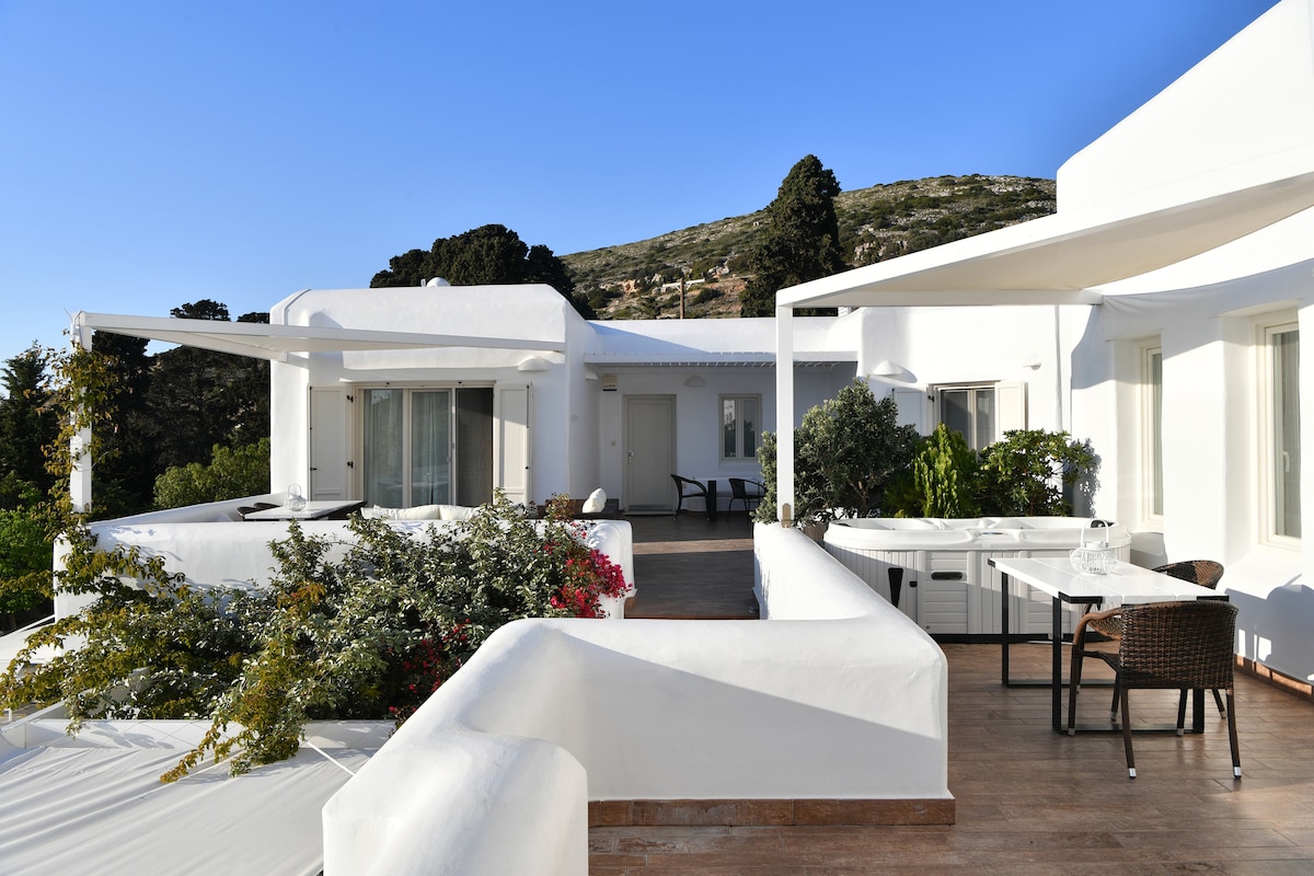 A spacious outdoor terrace is surrounded by lush greenery and vibrant flowers. Comfortable seating areas are visible, along with a dining table, all set against a backdrop of clean white architecture and clear blue skies.