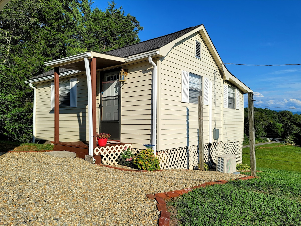 The exterior of a charming guest cottage is shown, featuring light-colored siding and a well-maintained landscape. A gravel pathway leads to the entrance, where a decorative lamp and potted flowers enhance the welcoming appearance. The structure is set against a backdrop of greenery beneath a clear blue sky.