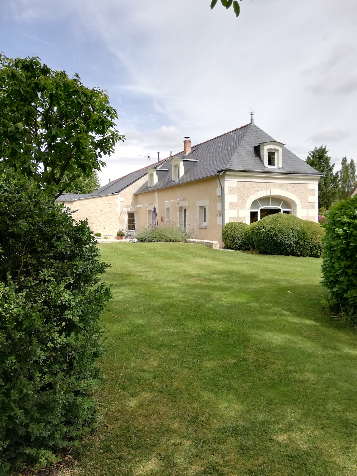 The exterior of a detached house is framed by well-maintained greenery, featuring a blend of stone and slate roofing. Expansive windows and an arched entrance complement the structure, which is surrounded by a lawn that offers ample open space in a tranquil setting.