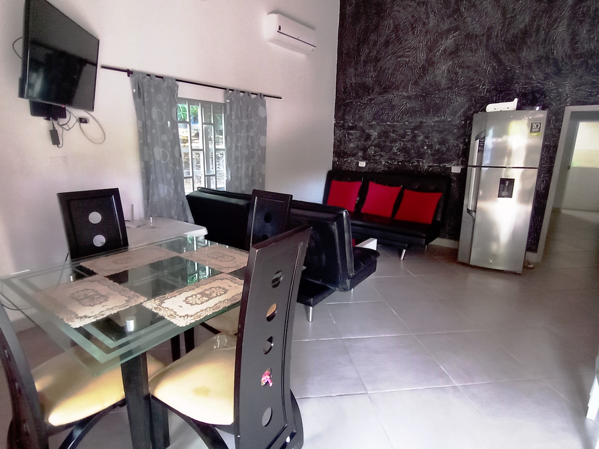 A living space features a glass dining table accompanied by four black chairs. A black sofa with red cushions is positioned against a textured wall. A flat-screen TV is mounted on the wall, and a silver refrigerator is seen in the background.