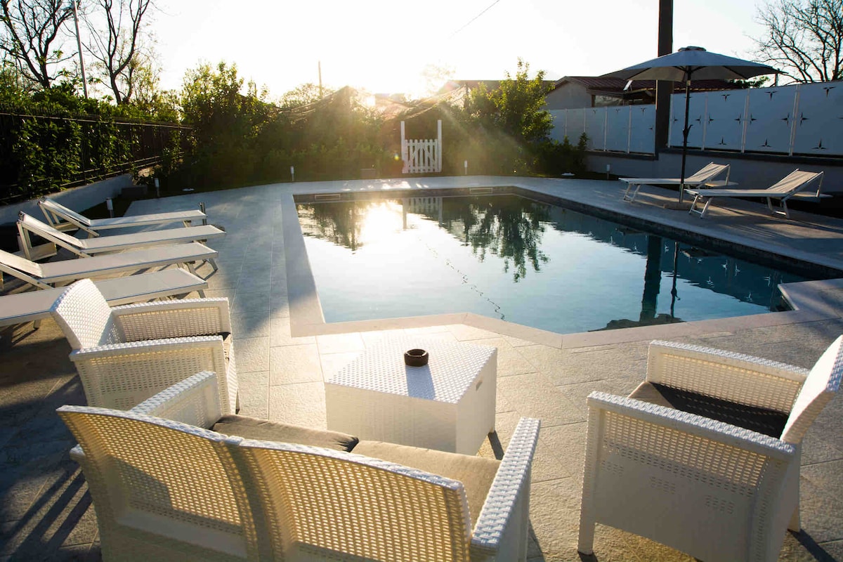 A private pool area is surrounded by neatly arranged white lounge chairs. Sunlight reflects off the clear water, creating a serene atmosphere. An umbrella provides shade, and a small table is positioned at the center of the seating arrangement.