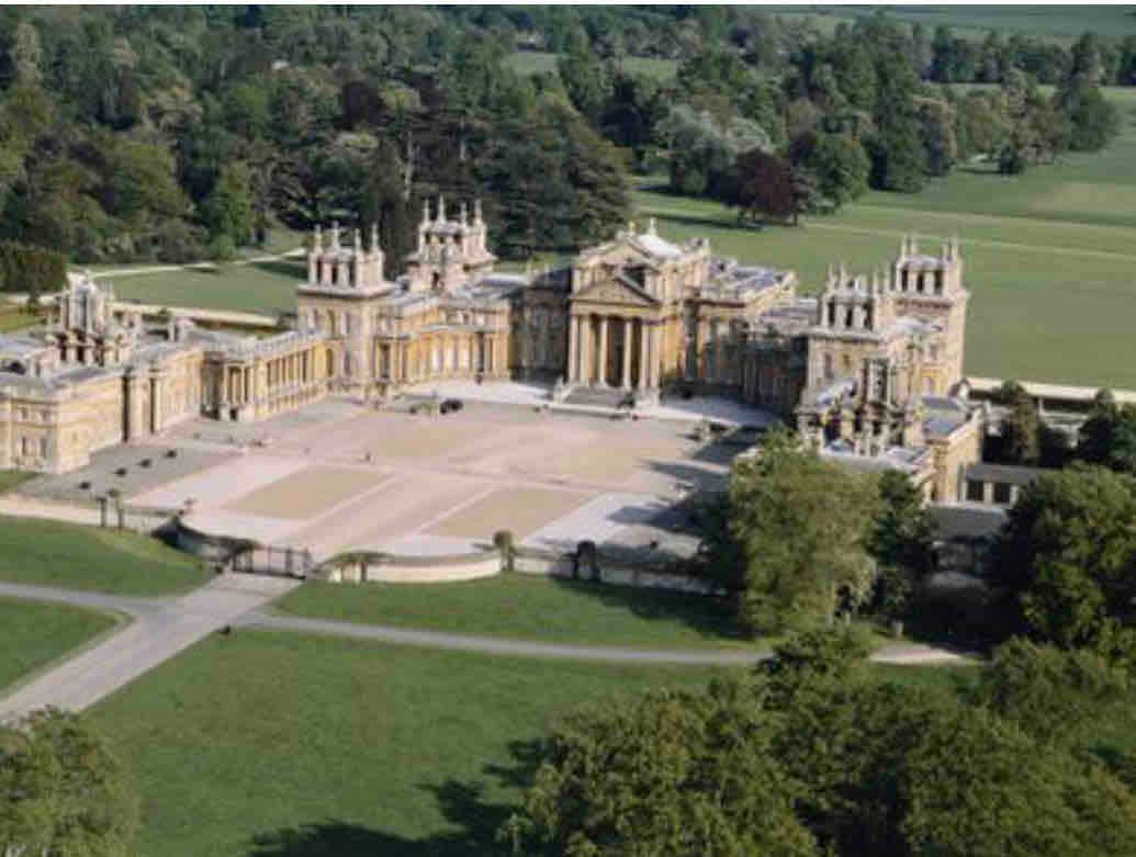 Aerial view of Blenheim Palace reveals its grand architecture and extensive landscaped gardens. The impressive structure features numerous towers and intricate stonework, surrounded by lush greenery. The expansive grounds include pathways and open areas that complement the historic building.