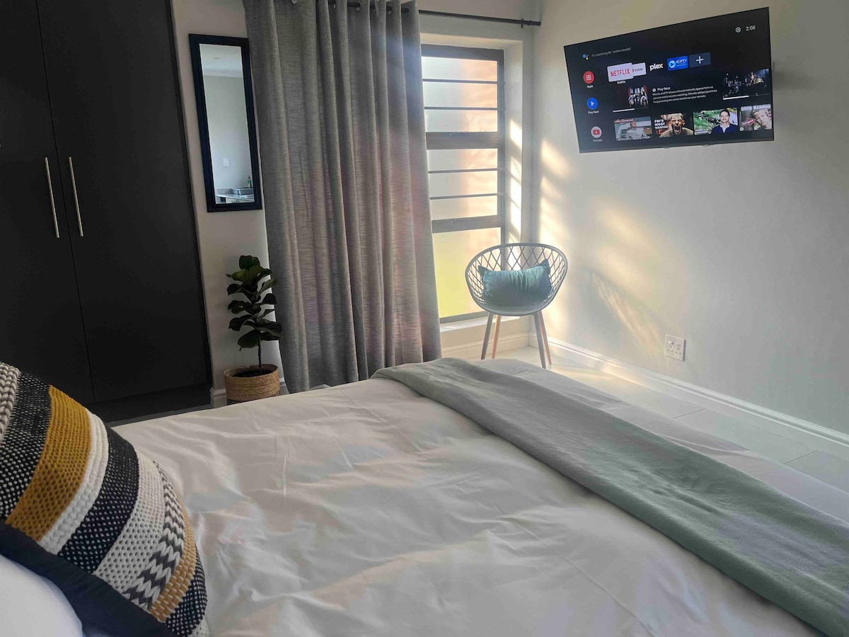 A spacious bedroom is highlighted with a large window allowing natural light to filter in through sheer curtains. A Smart TV is mounted on the wall across from the bed. A cozy accent chair and a small plant add warmth to the calming atmosphere.