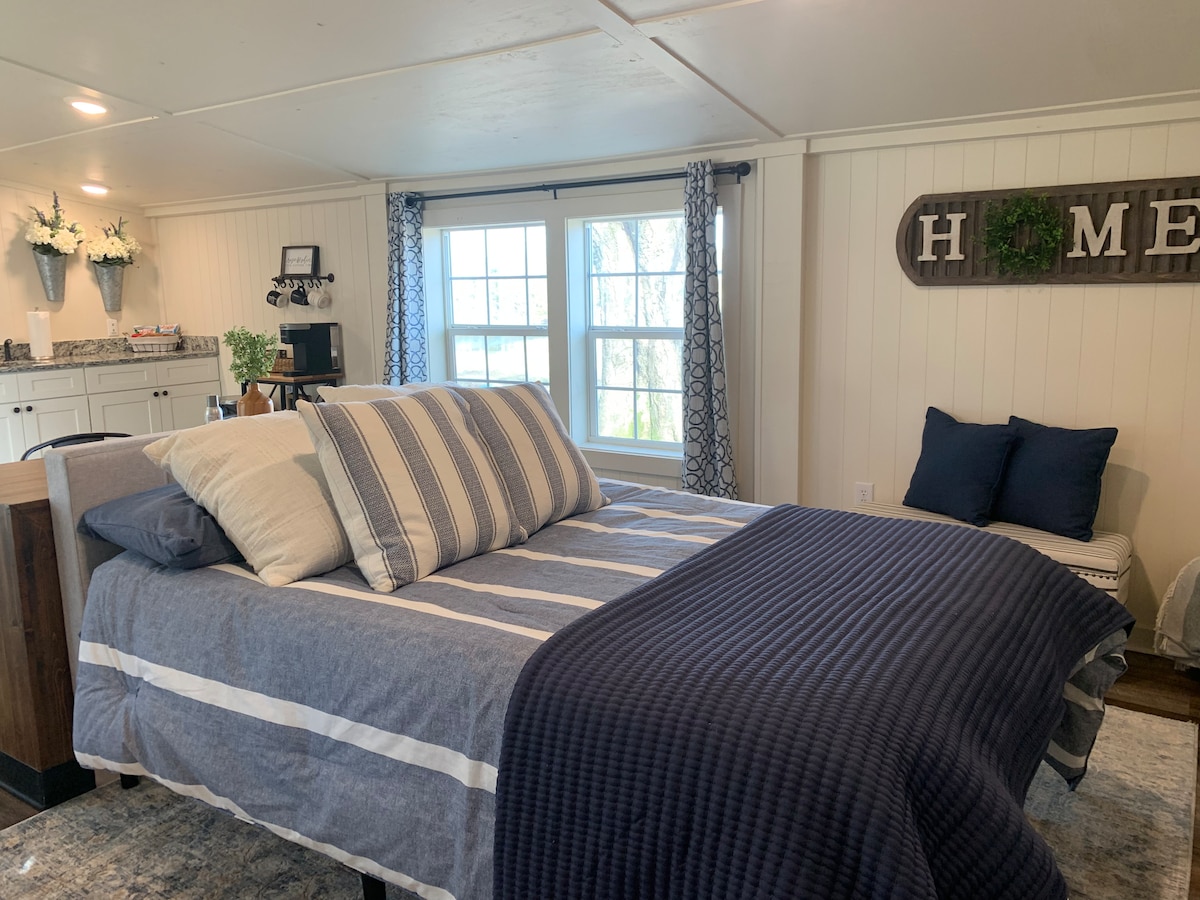 A spacious sleeping area features a neatly made bed with various patterned pillows and a textured blanket. A large window allows natural light to illuminate the room, while a cozy seating area and decorative elements add warmth to the space.