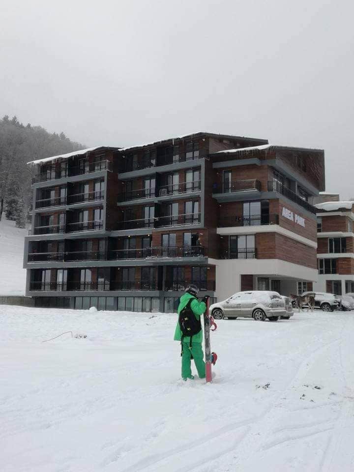 A modern ski lodge is visible, featuring large glass balconies and multiple levels. Snow blankets the ground, creating a serene winter landscape. A skier, dressed in bright green gear, stands in the foreground, preparing for a day on the slopes.