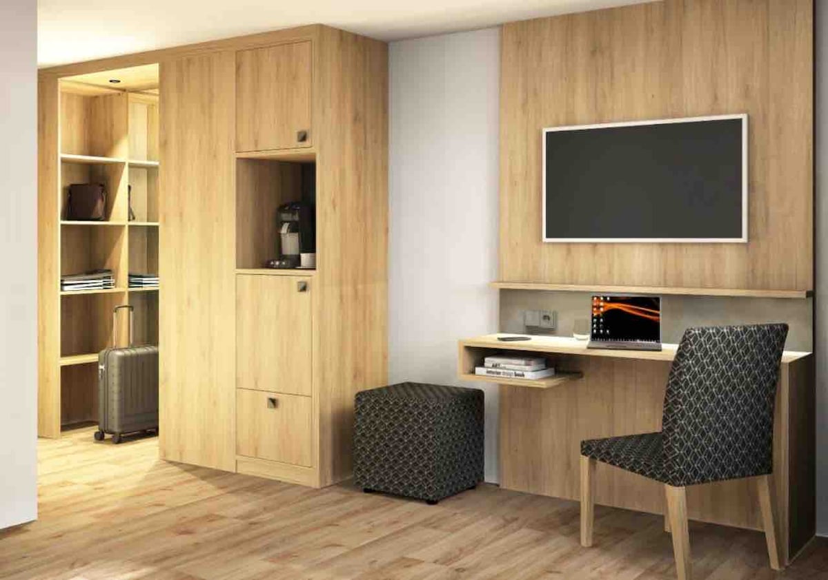 A modern room features light wooden cabinetry, providing ample storage options. A workstation with a sleek desk is positioned next to a comfortable chair. A flat-screen TV is mounted on the wall, and a cozy ottoman completes the inviting space.