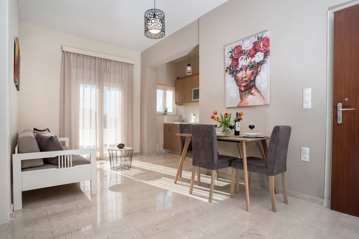 A bright and airy living space features a small dining area with a table and chairs, set against a backdrop of light-colored walls. Sunlight streams through large glass doors that lead to a balcony. A comfortable seating area, adorned with cushions, is positioned nearby.