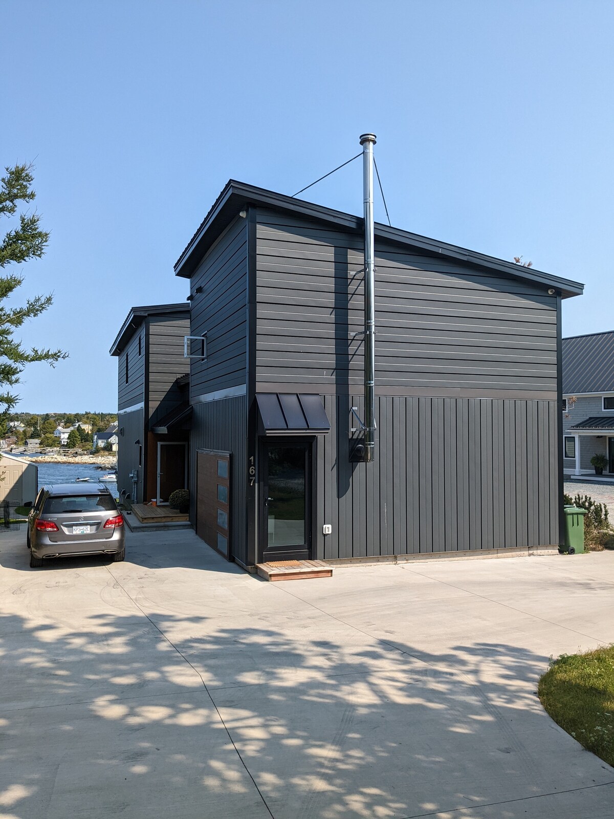 The exterior of a modern two-story cottage is depicted, showcasing a sleek dark wood facade with vertical paneling. A driveway leads to a side entrance, with a vehicle parked nearby. Sunlight casts shadows, highlighting the building's contemporary design.
