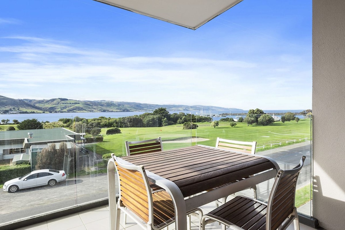 A dining table with wooden finishes is shown on a covered deck, surrounded by six chairs. Elevated views of green landscapes, the golf course, and the ocean are visible in the background. Clear blue skies enhance the serene environment.