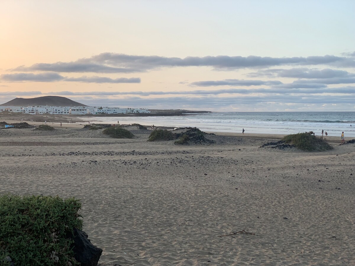 A serene beach scene is displayed, showcasing soft sandy dunes with sparse vegetation. Gentle waves are seen lapping at the shoreline, while a distant village with low white buildings is set against a backdrop of rolling hills. The sky reflects warm hues of evening light.